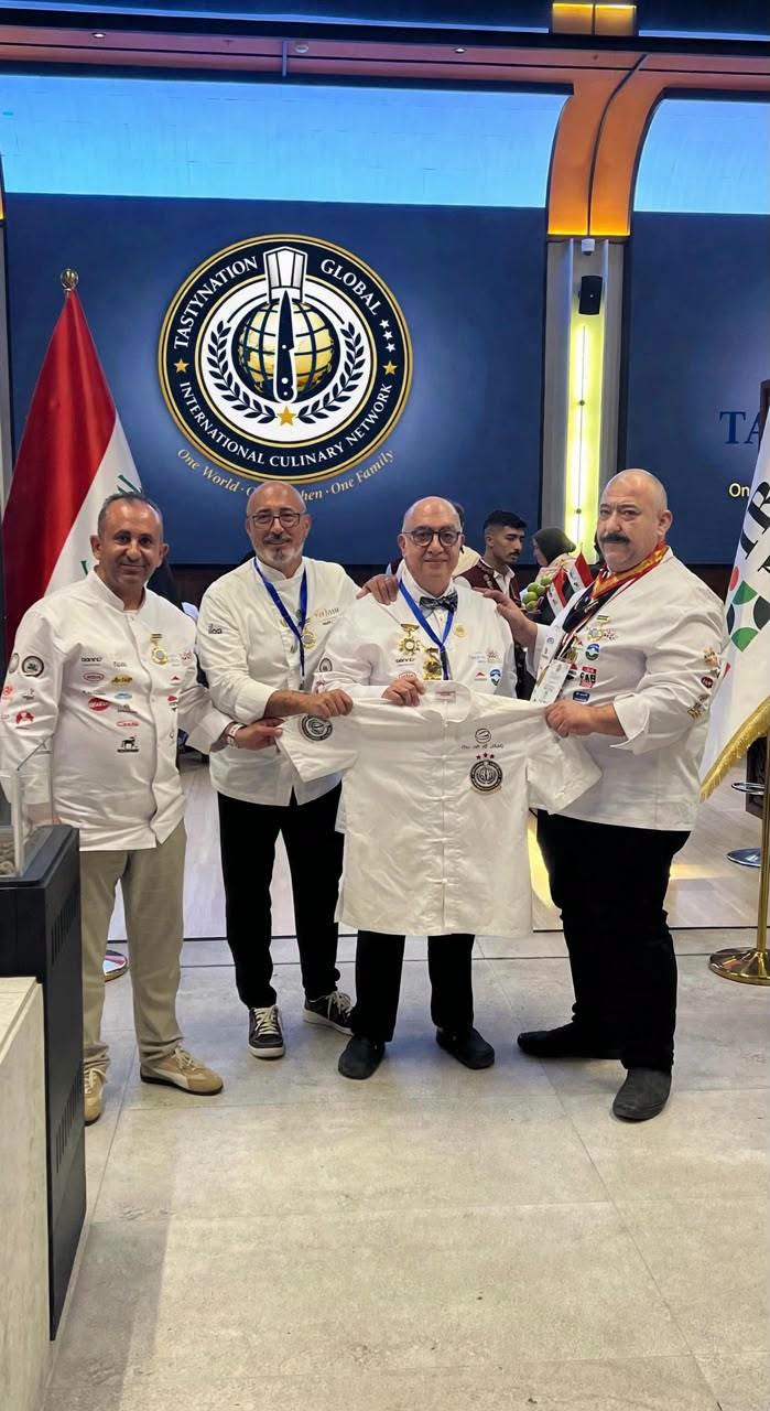 Tastynation Global representatives holding a chef jacket in front of the Tastynation Global emblem at the Iraq International Culinary Festival 2026